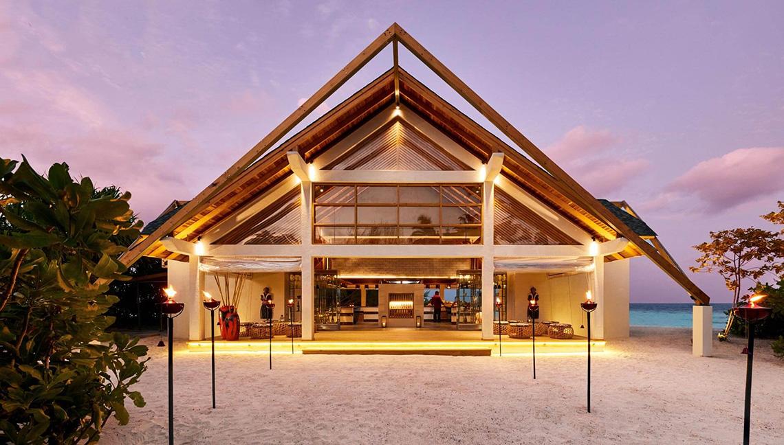 Außenansicht des Amari Havodda Maldives Hotels bei Nacht mit eingeschalteten Lichtern und Fackeln auf dem Strand.
