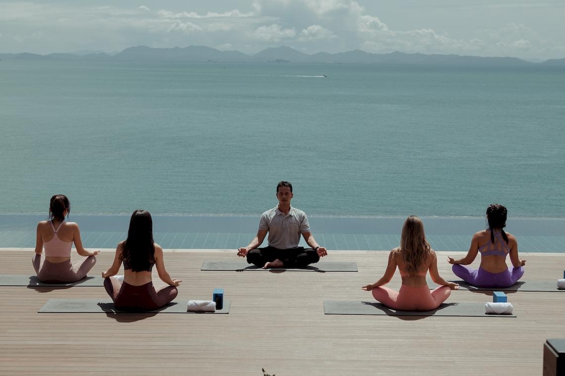 Ein Mann und vier Frauen machen Yoga auf einem Holzdeck am Strand mit Blick auf das Meer.