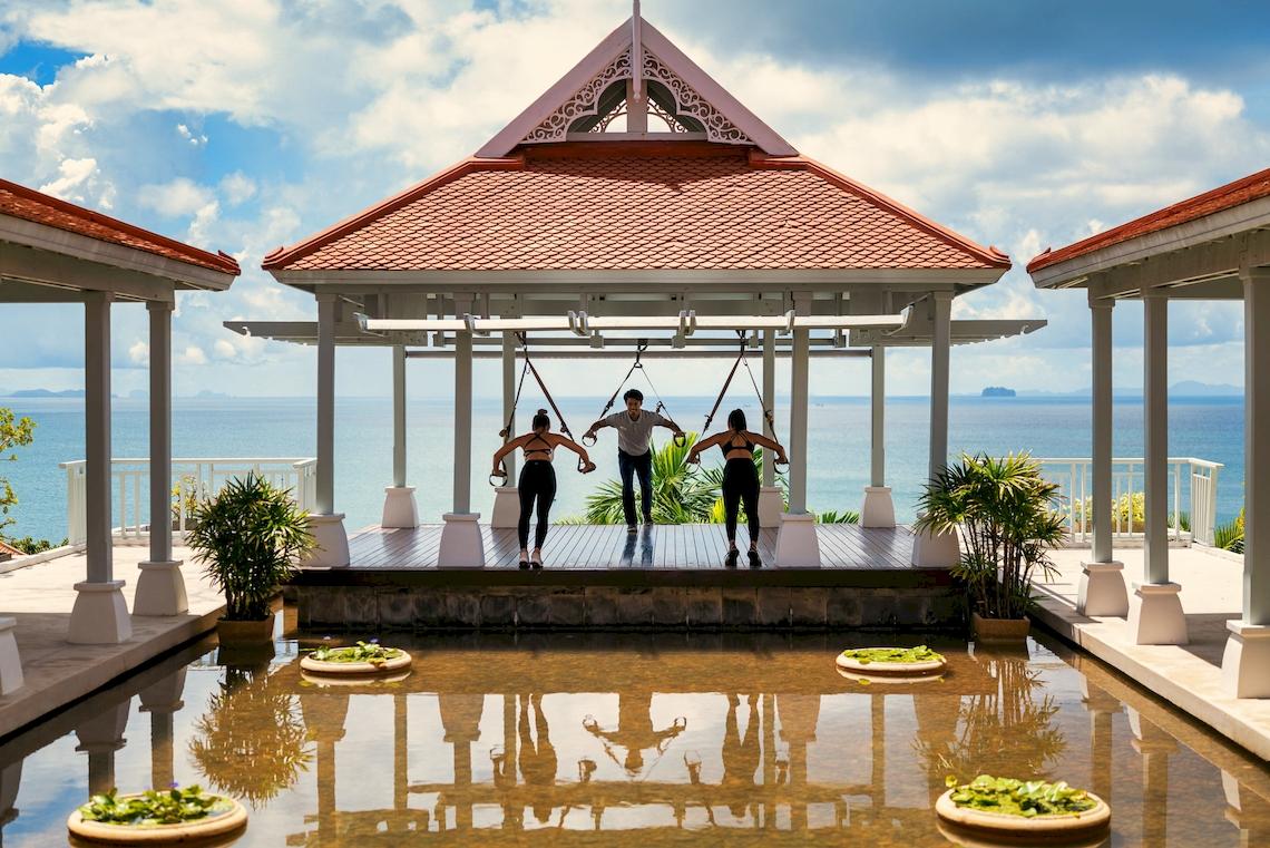 Drei Personen machen Yoga im Garten des Amatara Welleisure Resorts mit Blick auf das Meer.