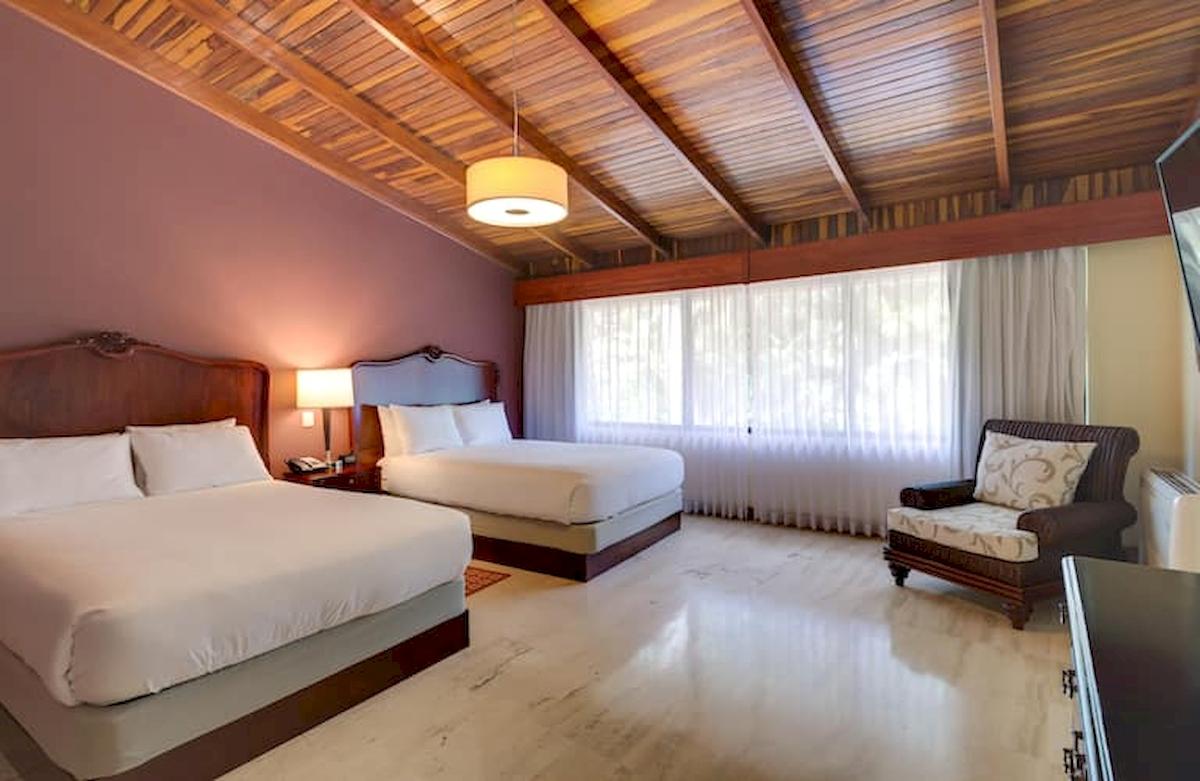 Doppelbett-Zimmer im DoubleTree by Hilton Hotel Cariari San Jose mit zwei Betten, Lampe, Fenstervorhängen und einem Sessel.