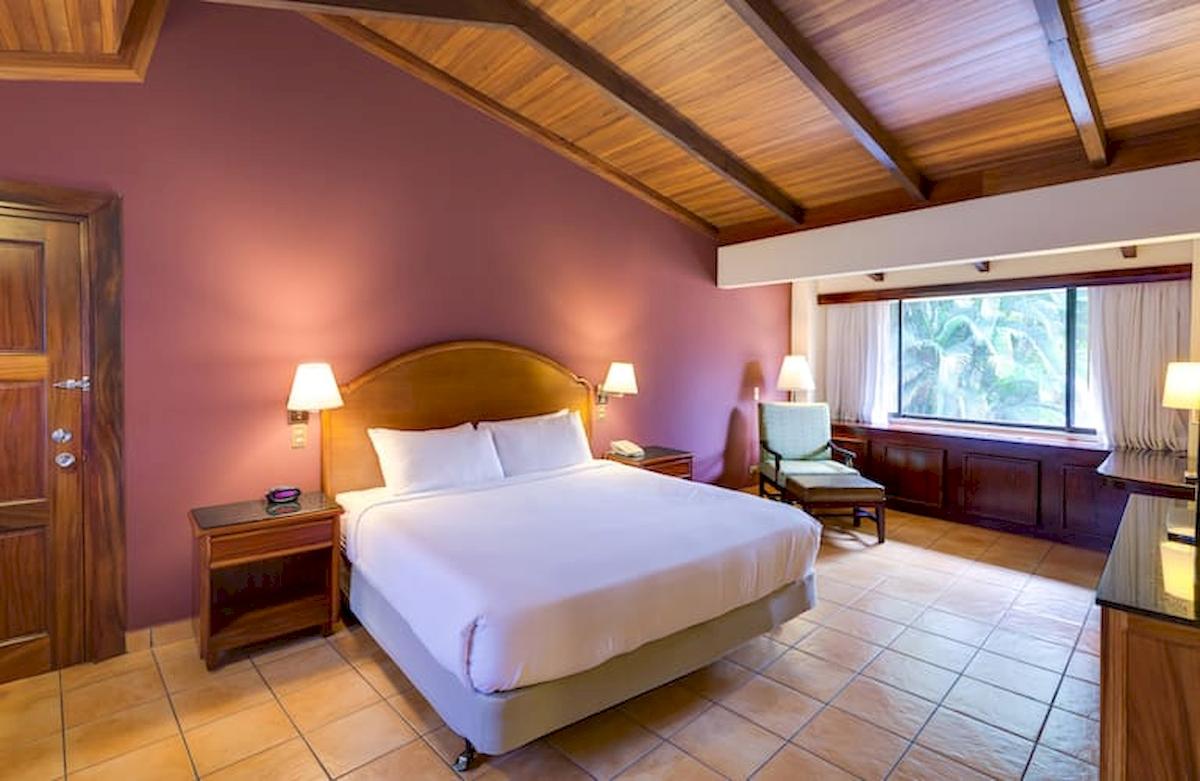 Doppelzimmer im DoubleTree by Hilton Hotel Cariari San Jose mit Bett, Nachttischen, Lampe, Fenster und Holzmöbeln.