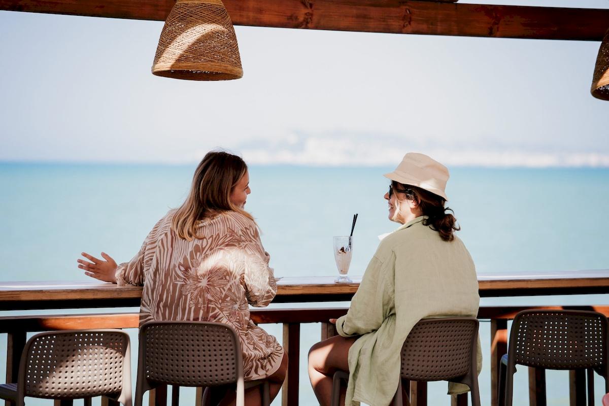 Zwei Frauen sitzen an einer Hotelbar mit Meerblick und unterhalten sich.