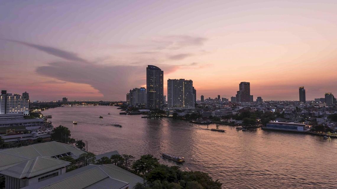 Aussicht auf Chatrium Hotel Riverside Bangkok bei Sonnenuntergang mit Fluss, Booten und Gebäuden