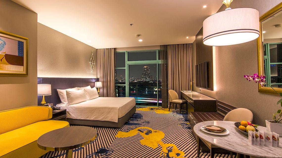 Innenansicht eines Zimmers im Chatrium Hotel Riverside Bangkok mit Bett, Couch, Schreibtisch und Aussicht auf die Stadt.