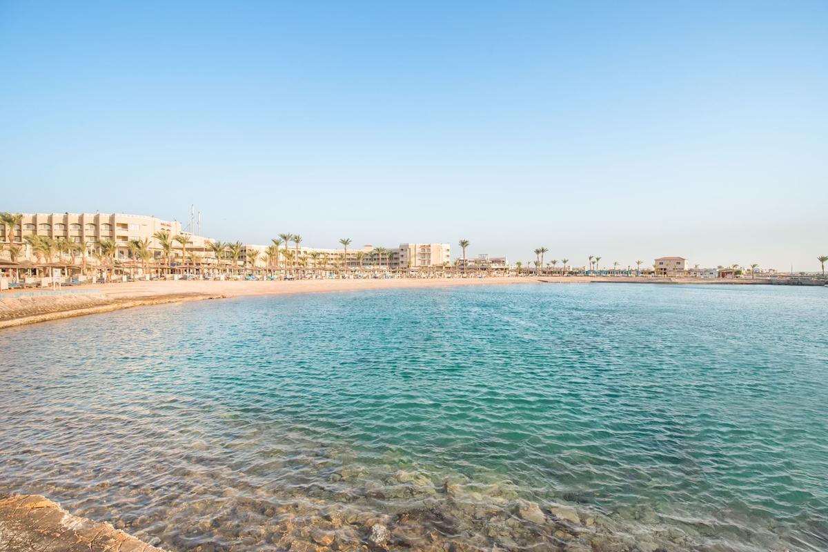 Strand des Pickalbatros Aqua Vista Resorts mit Gebäuden und Palmen im Hintergrund.