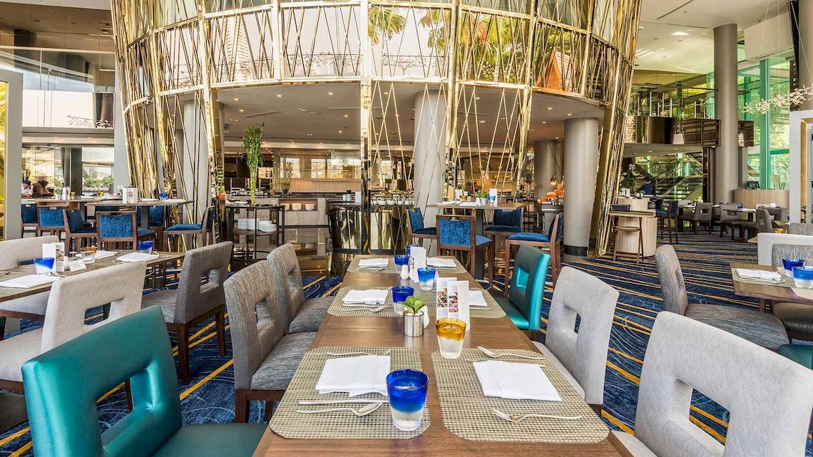 Restaurant-Innenbereich des Chatrium Hotel Riverside Bangkok mit langen Tischen, blauen Stühlen und einer zentralen goldenen Struktur.