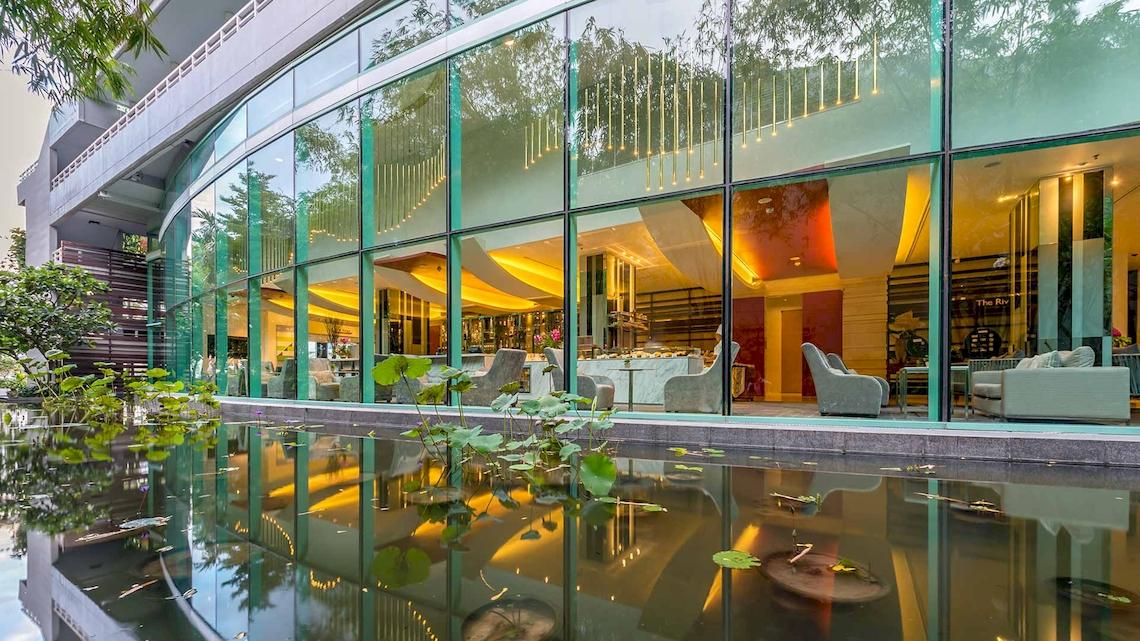 Außenansicht des Chatrium Hotel Riverside Bangkok mit Glasfassade und Wasserreflexion.
