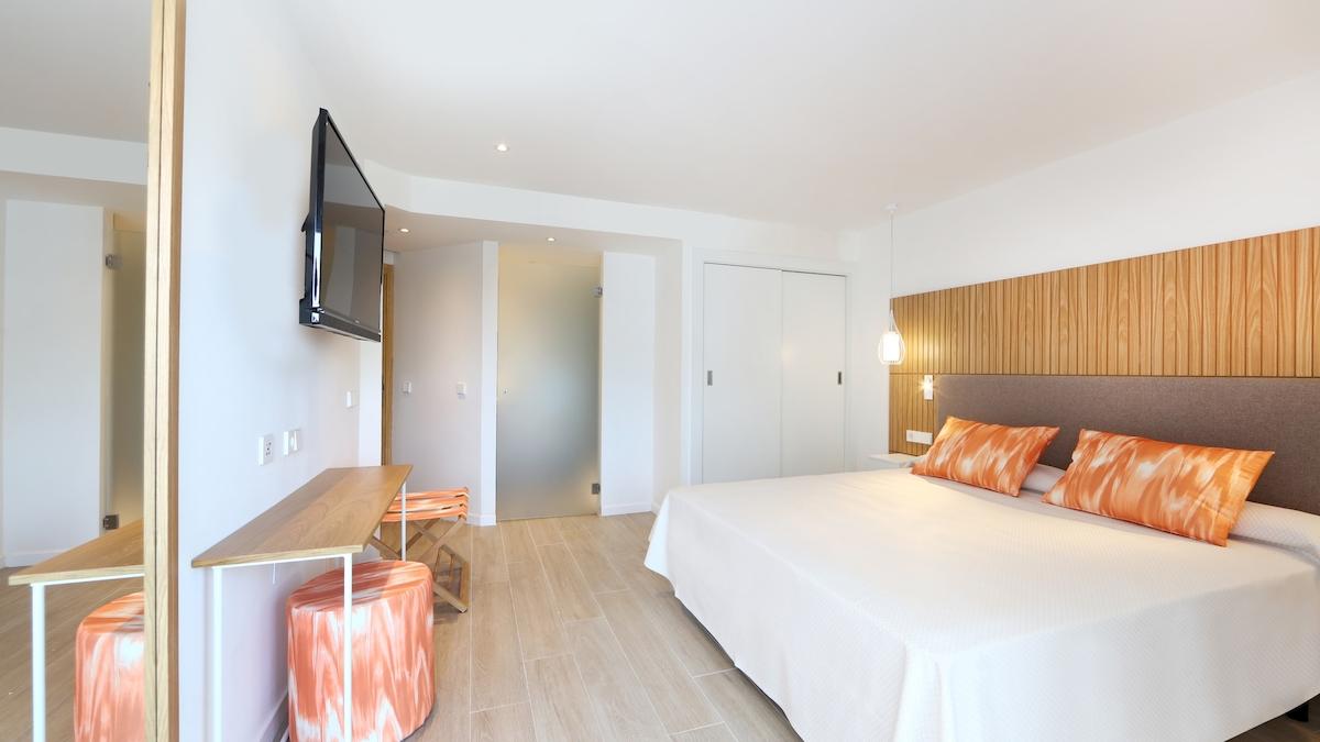 Innenansicht eines Schlafzimmers im Iberostar Playa de Muro Hotel mit weißem Bett, orangen Kissen und moderner Einrichtung.