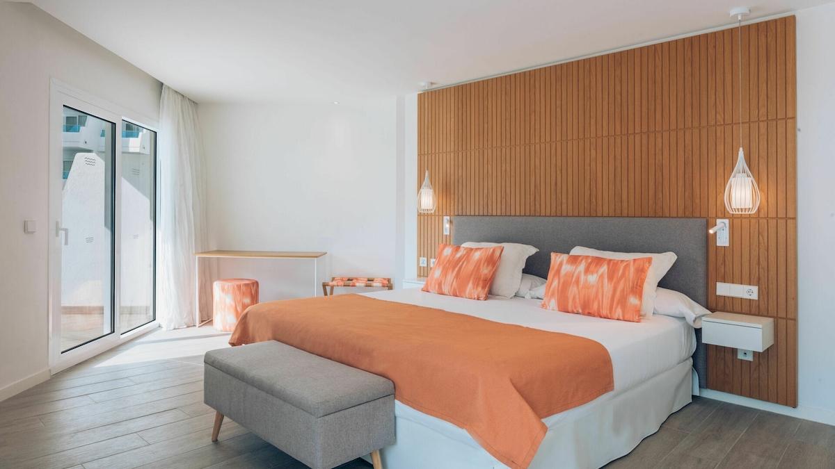 Schlafzimmer im Iberostar Playa de Muro Hotel mit Doppelbett, orange Bettwäsche, Nachttischen und begehbarer Dusche.