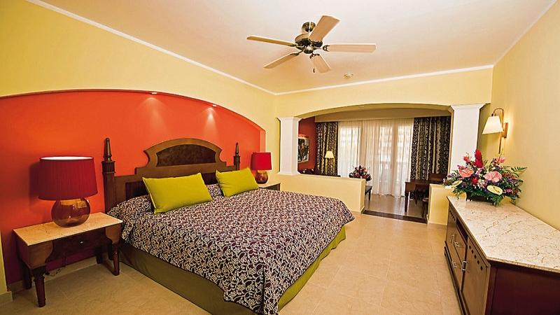 Schlafzimmer im Iberostar Selection Rose Hall Suites Hotel mit gelben und orangefarbenen Wänden, einem großen Bett, grünen Kissen und einem Deckenventilator.