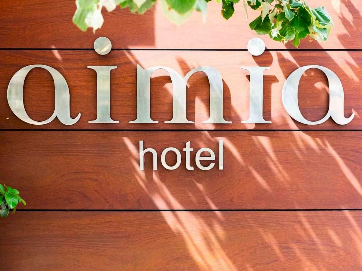 Schild des Aimia Hotels an einer braunen Holzwand mit Schatten und grünen Blättern