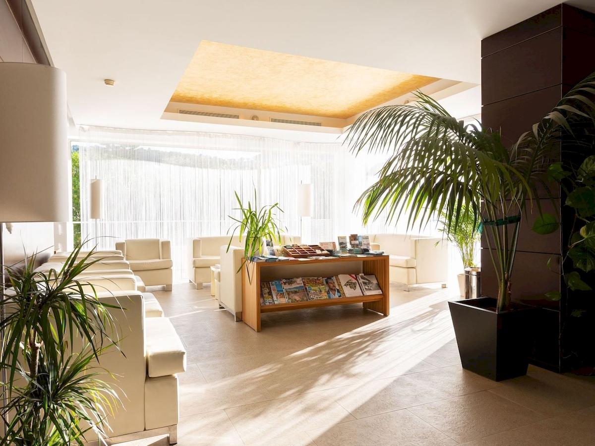 Empfangsbereich des Aimia Hotels mit weißen Sofas, Büchern auf dem Tisch und großen Pflanzen.