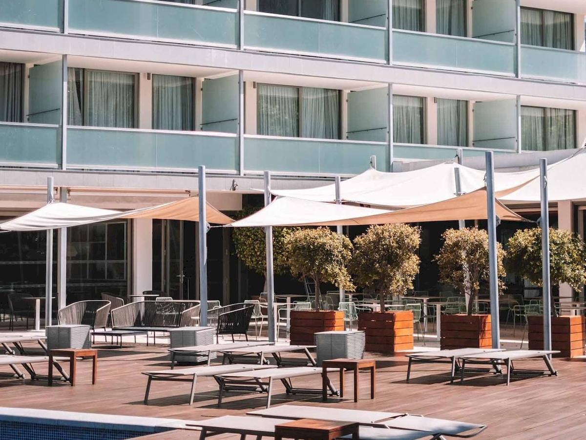 Außenbereich des Aimia Hotels mit Loungemöbeln, Sonnenschirmen und Bäumen.
