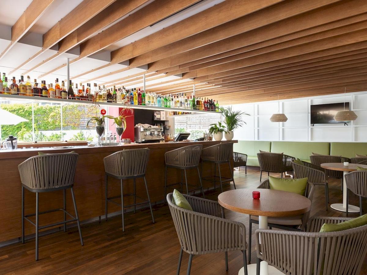 Innenansicht der Bar des Aimia Hotels mit Holzmöbeln, einer Theke und Pflanzen.