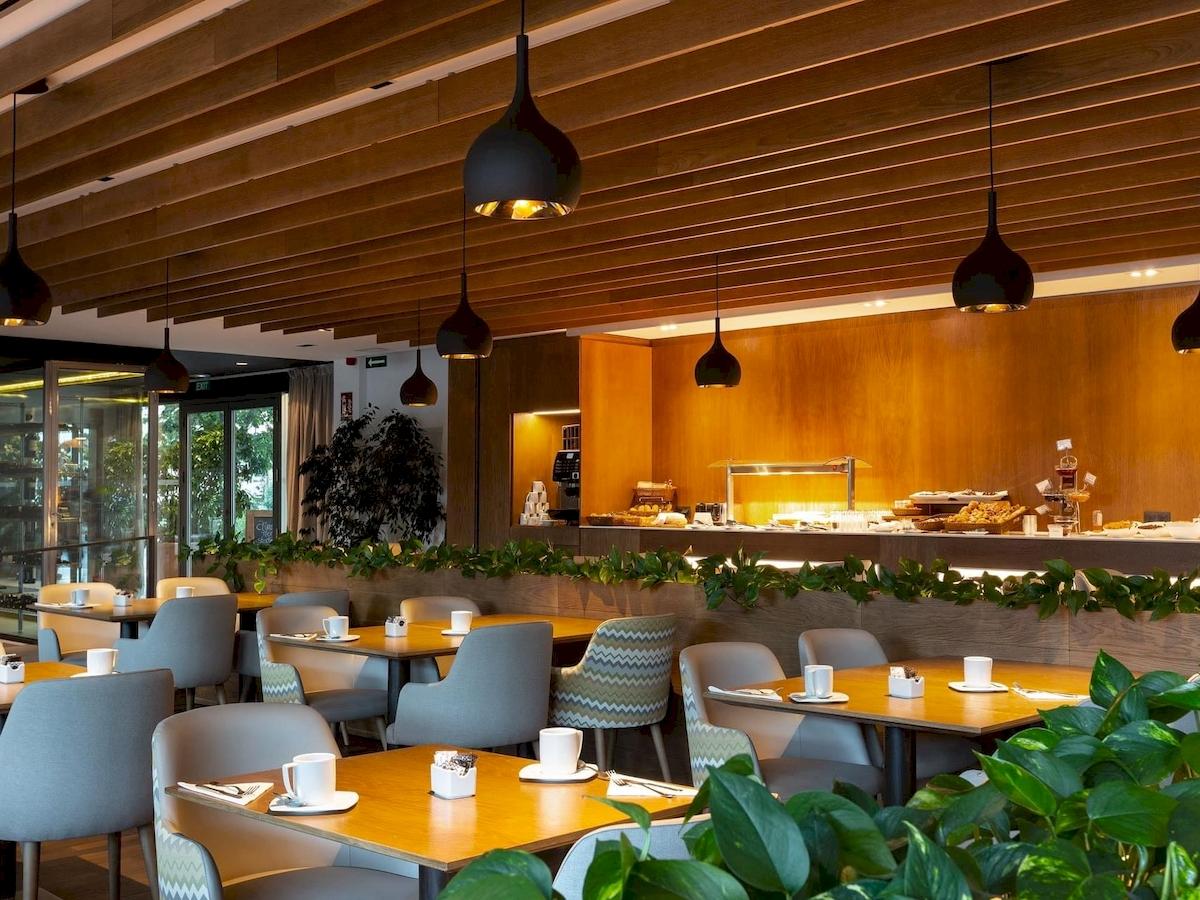 Frühstücksbereich des Aimia Hotels mit Tischen, Stühlen und Pflanzendekor.