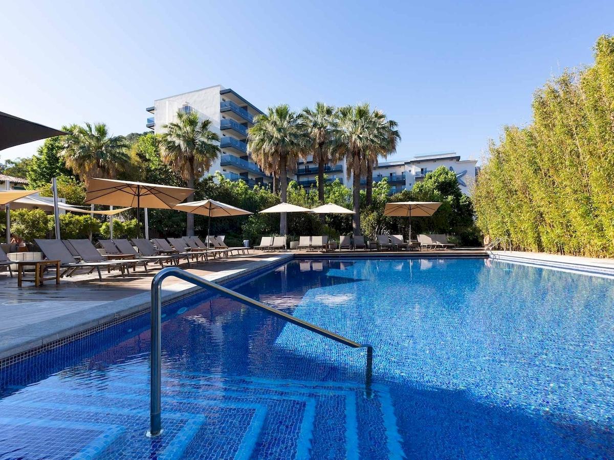 Außenpool des Aimia Hotels mit mehreren Liegen und Sonnenschirmen, umgeben von Bäumen und Gebäuden.