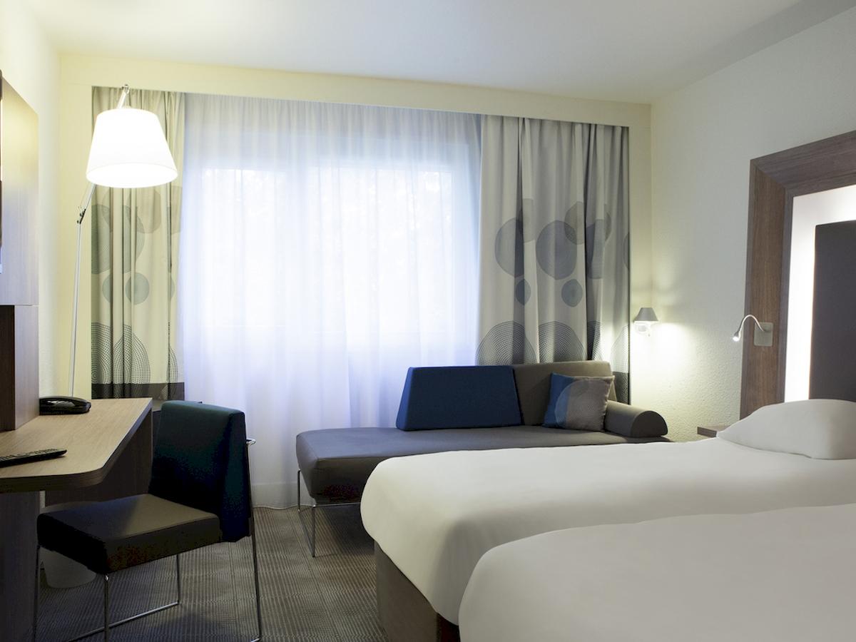 Innenansicht eines Hotelzimmers im Novotel London Canary Wharf mit Schreibtisch, Stuhl, Sofa und zwei Betten.