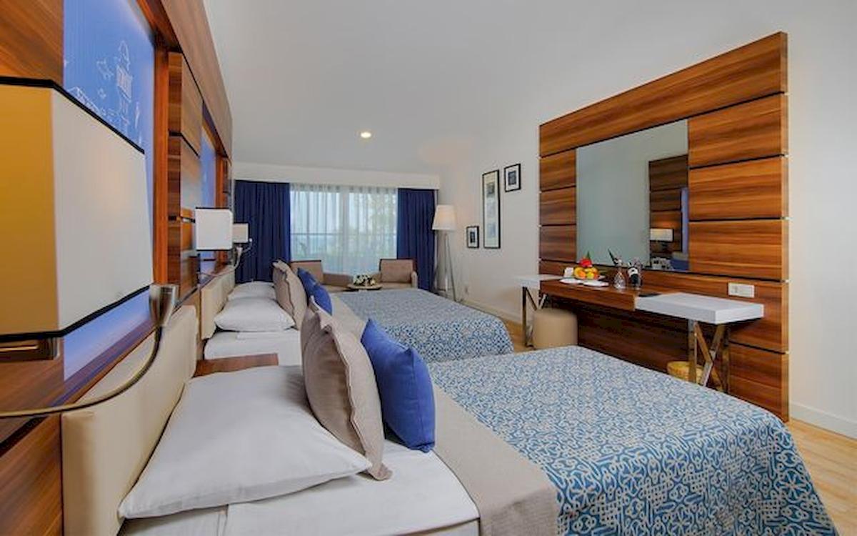 Zwei Doppelbetten in einem Zimmer im Limak Atlantis Deluxe Hotel & Resort mit blauen Vorhängen und Holzmöbeln.