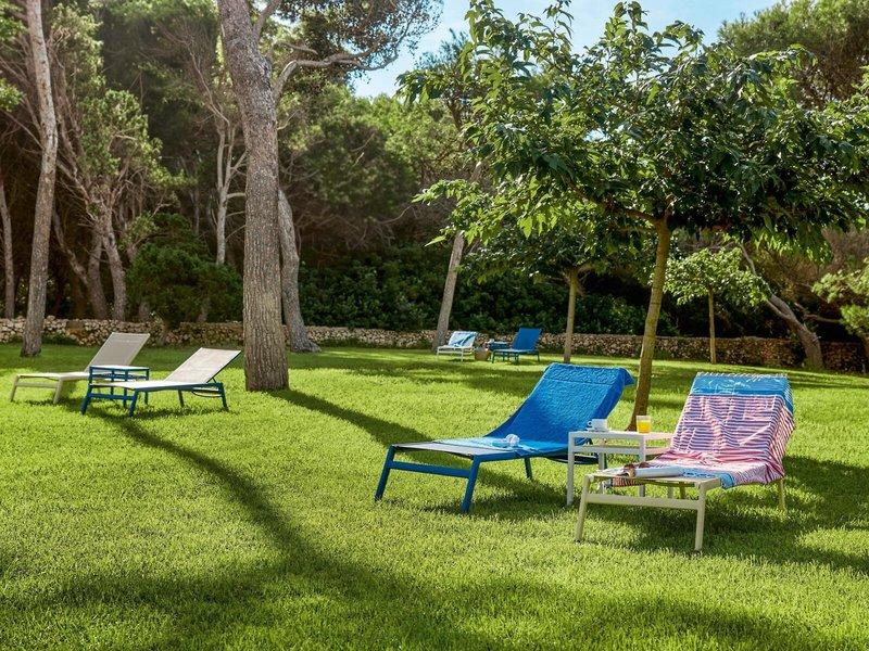 Gartenbereich des Victoria Menorca Hotels mit mehreren blauen und weißen Liegen auf grünem Rasen.