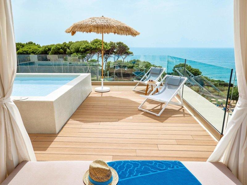 Terrasse mit Meerblick im Victoria Menorca Hotel, mit Jacuzzi, Sonnenliegen und Sonnenschirm.