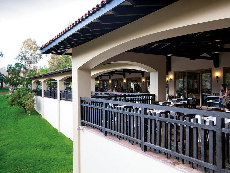 Restaurantbalkon im Melas Holiday Village mit grüner Rasenfläche und bestuhlten Tischen.