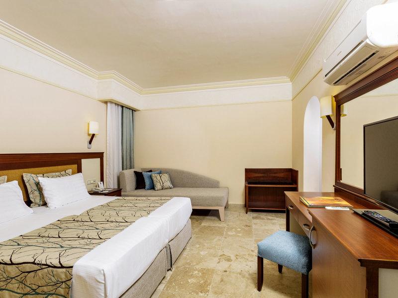Hotelzimmer im Melas Holiday Village mit Bett, Sofa, Schreibtisch und Fernseher.