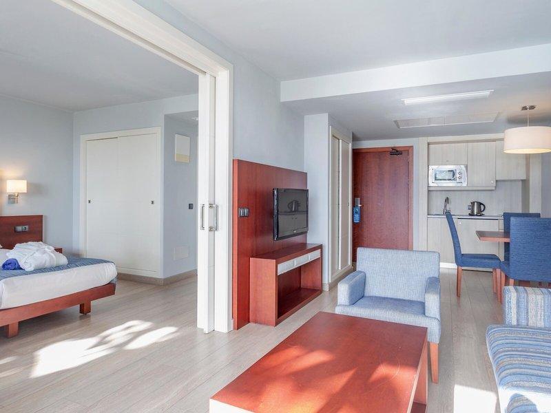 Innenansicht eines Zimmers im Marins Playa Suites Hotel mit Bett, Fernseher, Couch, Esstisch und Küchenbereich.
