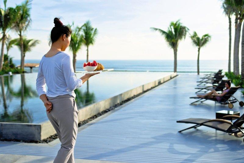 Frau des TIA Wellness Resorts mit Tablett am Pool, Blick auf das Meer und Lounge-Stühle