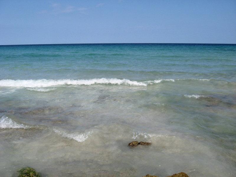 Klares Meer mit sanften Wellen am Strand und einigen Felsen im Vordergrund.