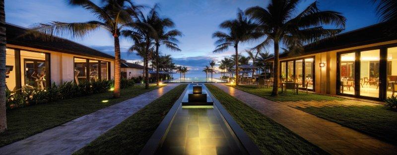 Nachtansicht des TIA Wellness Resorts mit einem langen Wasserlauf und beleuchteten Palmen.