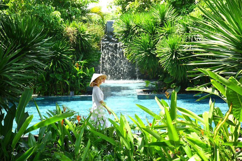 Eine Frau im weißen Kleid und Hut steht im TIA Wellness Resort Garten neben einem Pool und Wasserfall.