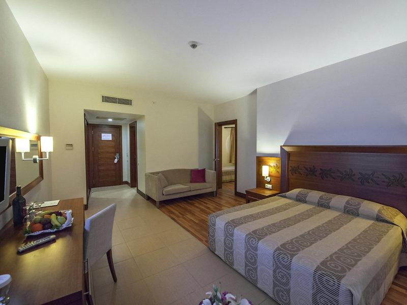 Hotelzimmer im Kirman Leodikya Resort mit Doppelbett, Sofa, Schreibtisch und Obstplatte.