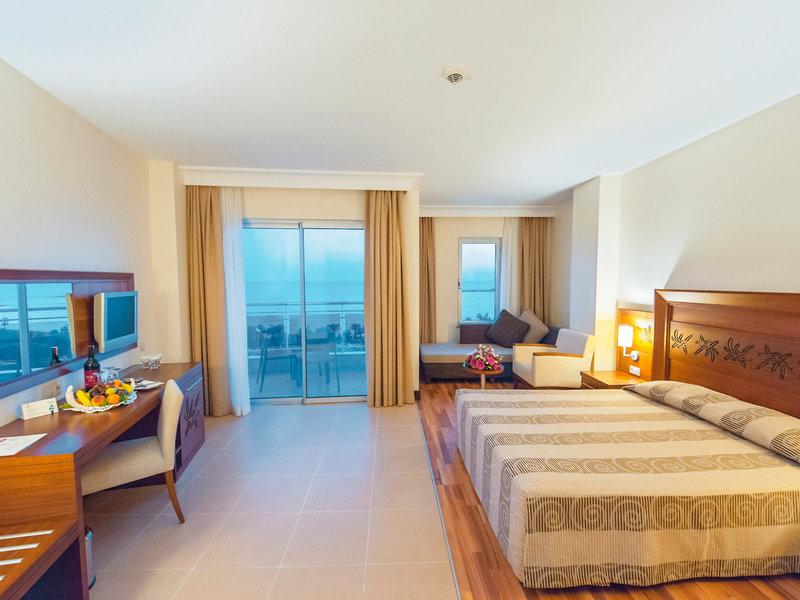 Hotelzimmer im Kirman Leodikya Resort mit großem Bett, Schreibtisch und Blick auf die Terrasse.