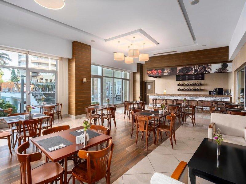 Innenansicht des Restaurants im Kirman Leodikya Resort mit Tischen, Stühlen und Blick auf eine Terrasse.