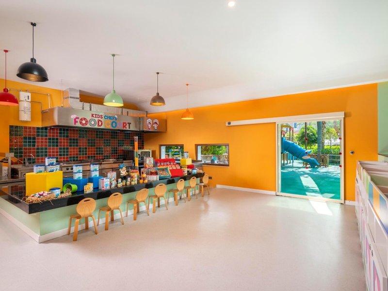 Kinderkoch-Essensbereich im Kirman Leodikya Resort mit gelben Wänden, Holzstühlen und Spielplatz im Hintergrund.