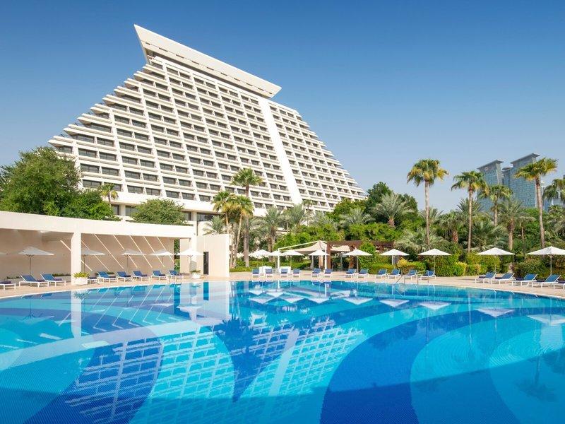 Ein Swimmingpool vor dem Sheraton Doha Resort Convention Hotel mit Sonnenliegen und Sonnenschirmen.