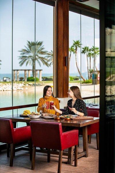 Zwei Frauen im Sheraton Doha Resort Convention Hotel, sitzend am Tisch mit Essen und Getränken vor einer Panorama-Seeansicht.