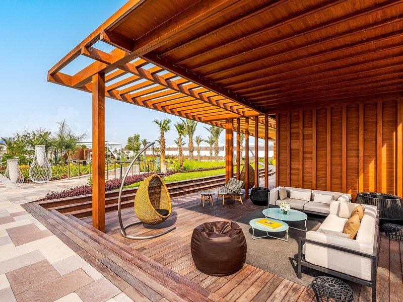 Terrasse des Rixos Gulf Hotel Doha mit hölzernem Überdach, Sitzmöbeln und Blick auf einen Garten