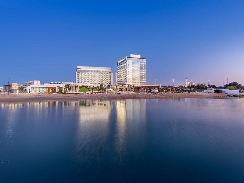Luftaufnahme des Rixos Gulf Hotel Doha am Strand mit reflektierten Gebäuden im Wasser.