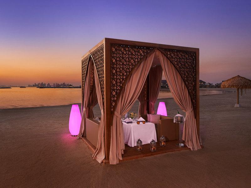 Strand-Gazebo mit Tisch und Sesseln, umgeben von pinken Leuchten und Vorhängen, bei Sonnenuntergang.