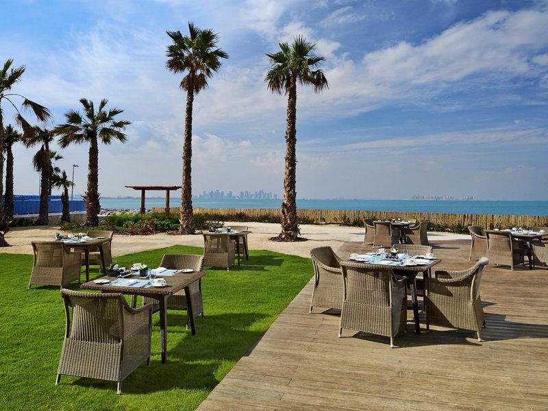 Außenbereich des Banana Island Resort Doha by Anantara mit Tischen, Stühlen und Palmen, Blick aufs Meer.