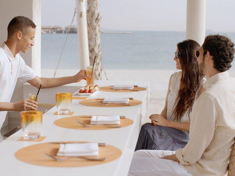 Drei Gäste sitzen am Tisch am Strand des Banana Island Resorts Doha mit Getränken und Blick aufs Meer.