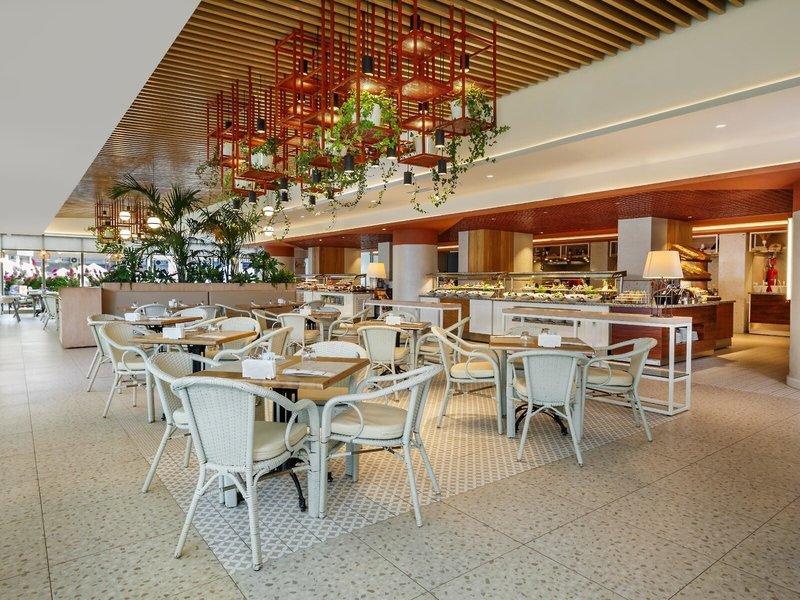 Innenbereich des Grand Park Lara Hotels mit geometrischen Mustern, Tischen und Stühlen, hängenden Pflanzen und einem Büffet.