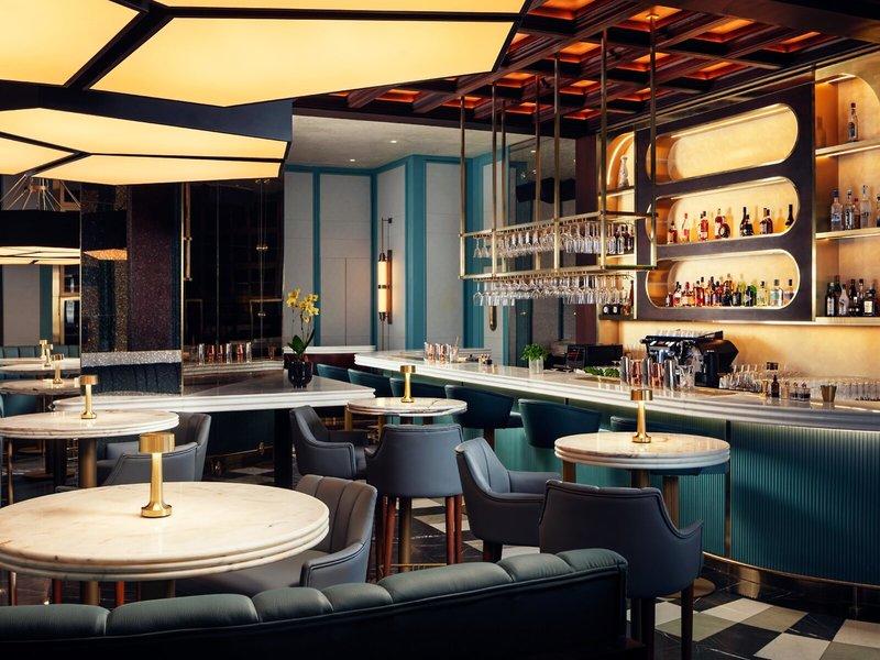Innenansicht der Bar im St. Regis Al Mouj Muscat Resort mit Marmortischen, Stühlen und einer Theke mit vielen Flaschen.