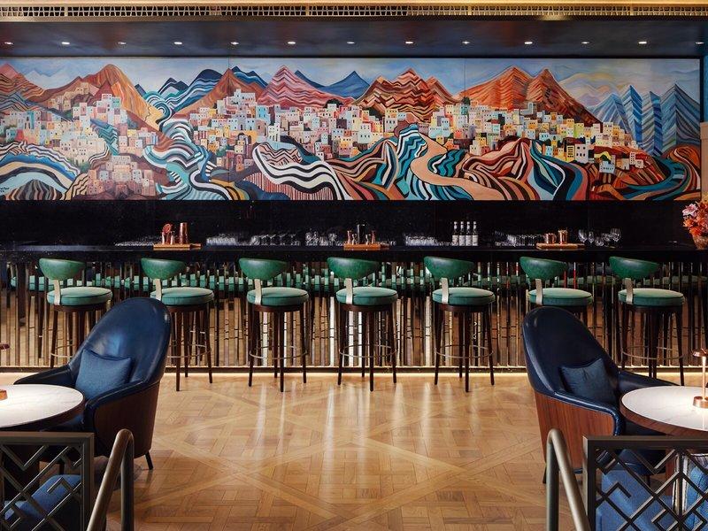 Innenansicht der Bar im St. Regis Al Mouj Muscat Resort mit buntem Wandgemälde und grünen Barhockern.