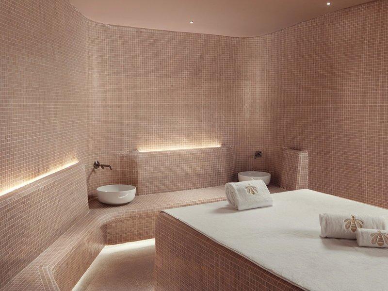 Spa-Behandlungsraum im St. Regis Al Mouj Muscat Resort mit gefliesten Wänden, Sitzbänken, weißen Waschbecken und Handtüchern.