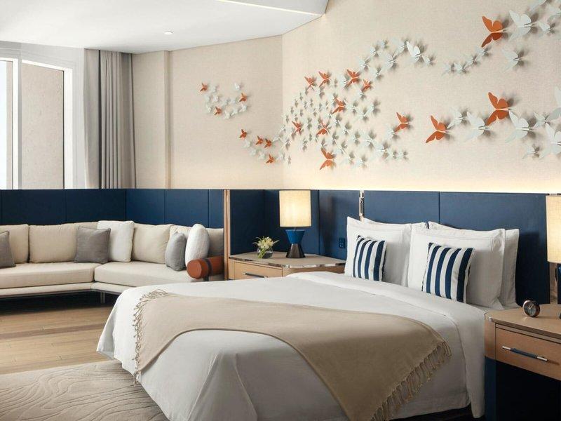 Schlafzimmer im St. Regis Al Mouj Muscat Resort mit großem Bett, Sofa, Nachttischen und blauen Kissen.