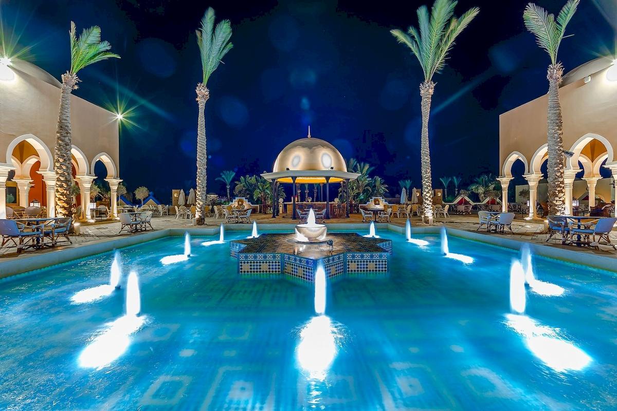 Nachtansicht des Swimmingpools im Grand Makadi Hotel mit Wasserfontaene und umliegenden Liegestuehlen.