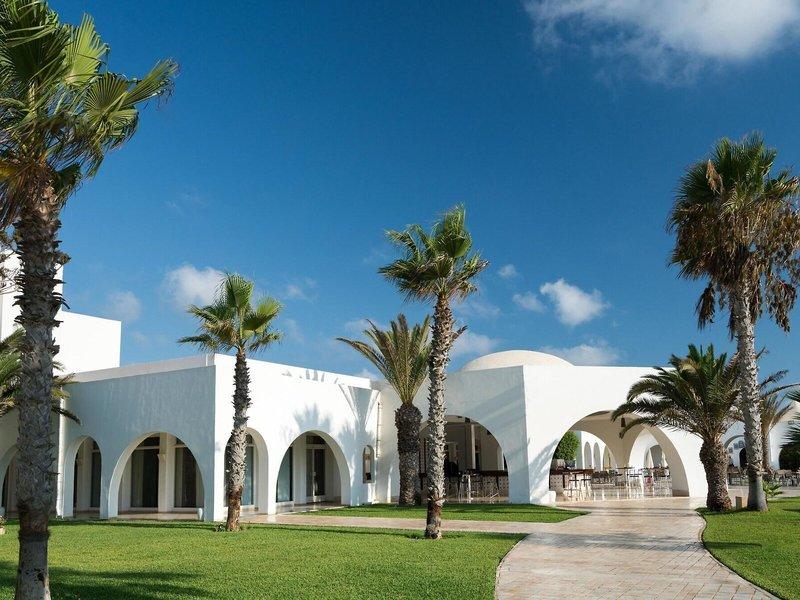 Außenansicht des weißen Hotels Iliade Djerba by Magic Hotels mit Palmen und Rasen