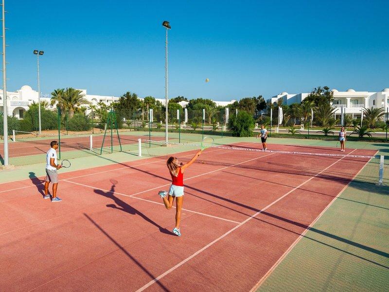 Tennisplatz im Hotel Iliade Djerba mit Spielern und Gebäuden im Hintergrund.